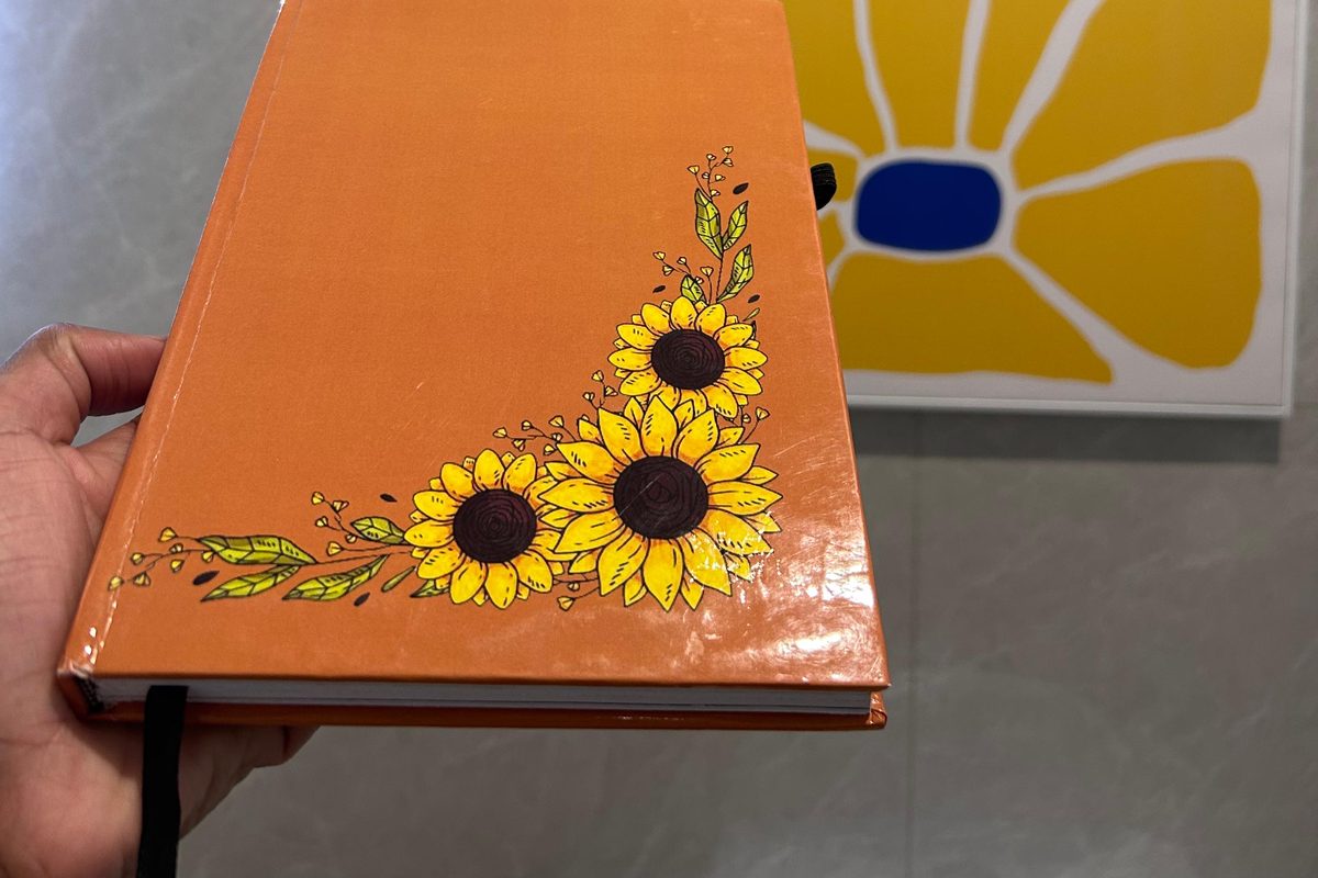 An orange hardback journal, hand-painted with sunflowers, held in one hand.